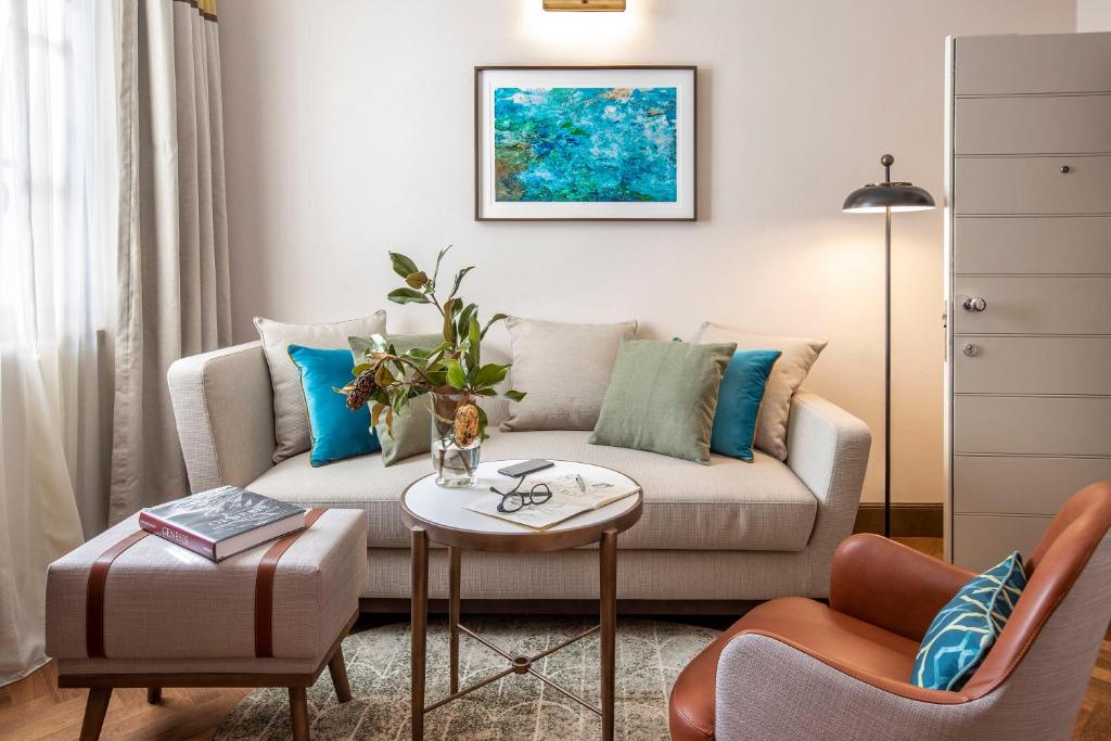 Mūsdienīgs viesistabas interjers ar ērtu dīvānu, dekoratīviem spilveniem, ziedu kompozīciju un mākslas darbu uz sienas. Telpā ir arī kafijas galdiņš un apgaismojums, radot mājīgu atmosfēru.