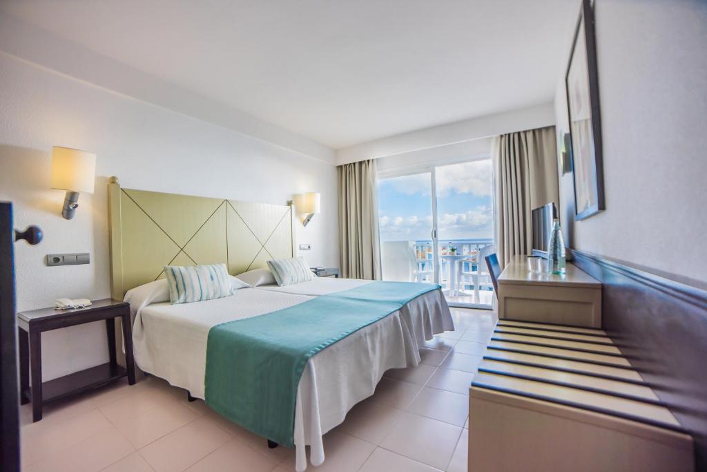 Mūsdienīga viesnīcas numura interjers ar divām gultām, zilu gultas pārklāju un plašu logu, kas piedāvā skatu uz āra terasi. Gaismas toņi un minimālistisks dizains rada relaksējošu atmosfēru.