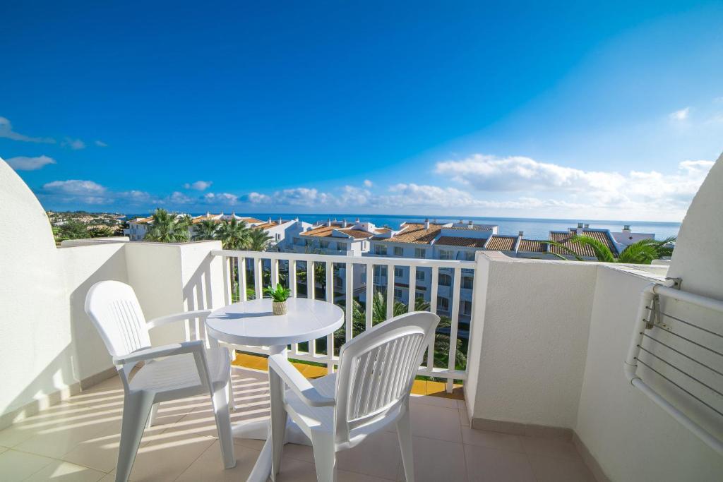 Balkons ar divām baltām krēsliem un galdu, no kura paveras skats uz jūru un apkārtējiem mājokļiem zem saulainām debesīm.
