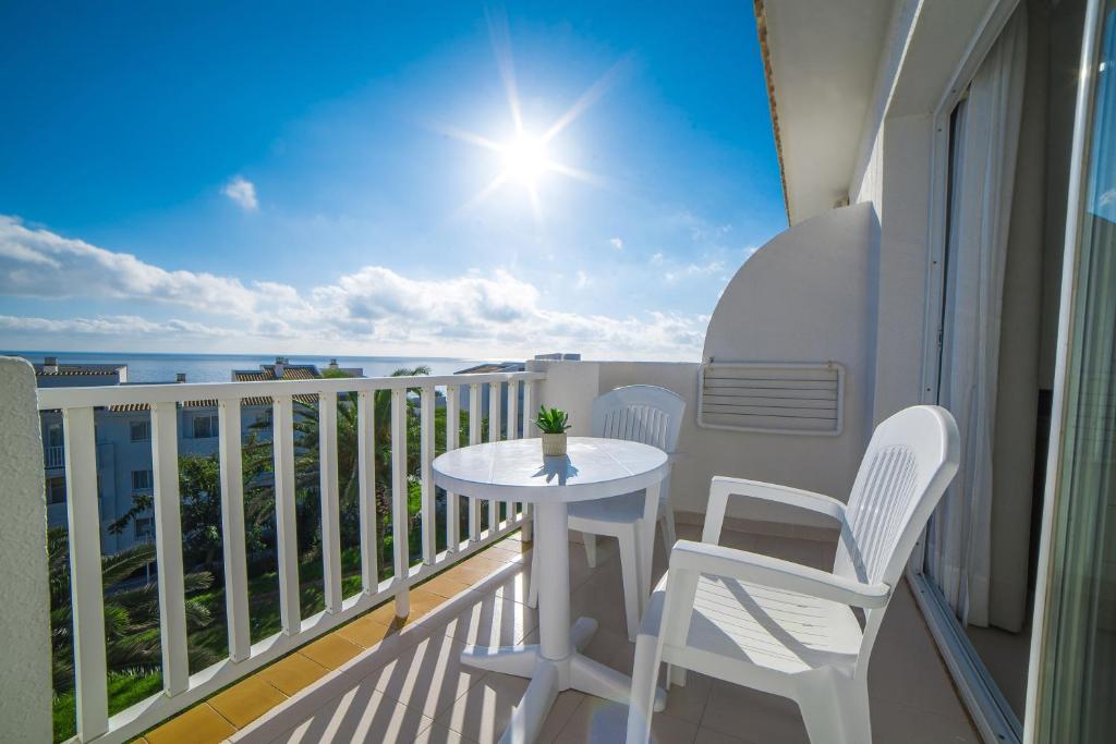 Saulaina balkona skats ar baltu galdu un krēsliem, kurā ir augs. Fons ir debesis ar mākoņiem un jūras skats.