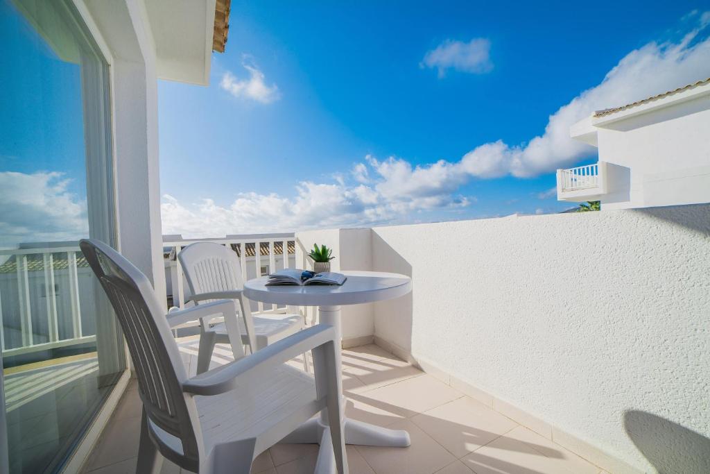 Saulaina terase ar baltu galdu un krēsliem, grāmata uz galda un zaļš augs. Aiz terases redzams skaidrs debesis ar dažiem mākoņiem.