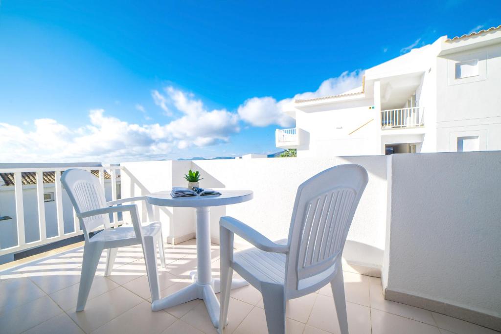 Balkons ar baltām plastmasas mēbelēm, uz galda atvērta grāmata un neliela auga pods, fons ar zilu debesu un mākoņu ainavu.