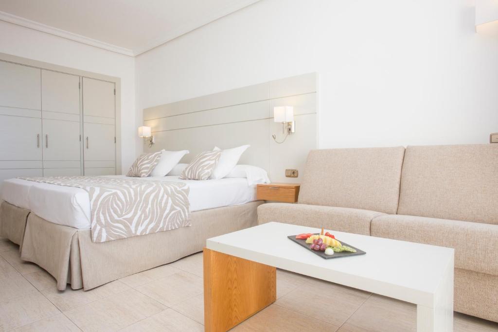 Mūsdienīgs viesnīcas numurs ar divguļamo gultu, dīvānu un elegantu galdu, uz kura atrodas augļu plate. Gaismas krāsas un vienkāršs dizains rada patīkamu atmosfēru.