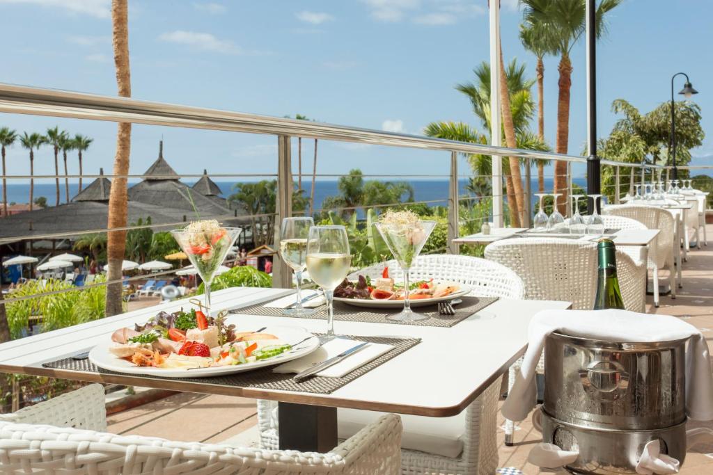 Eleganta restorāna terase ar skatu uz jūru, kurā pasniegti izsmalcināti ēdieni un dzērieni. Atpūtas zona ar palmām un peldbaseiniem rada relaksējošu atmosfēru.