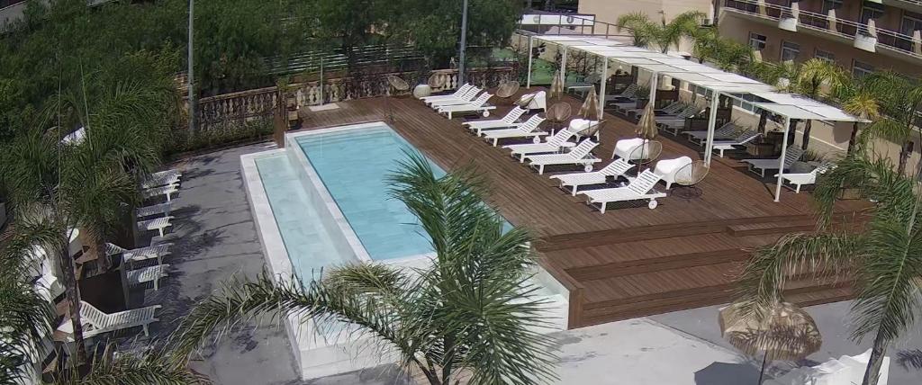 Peldbaseins ar saulessargiem un atpūtas krēsliem, apkārt palmām un viesnīcas ēkai, radot relaksējošu atmosfēru.