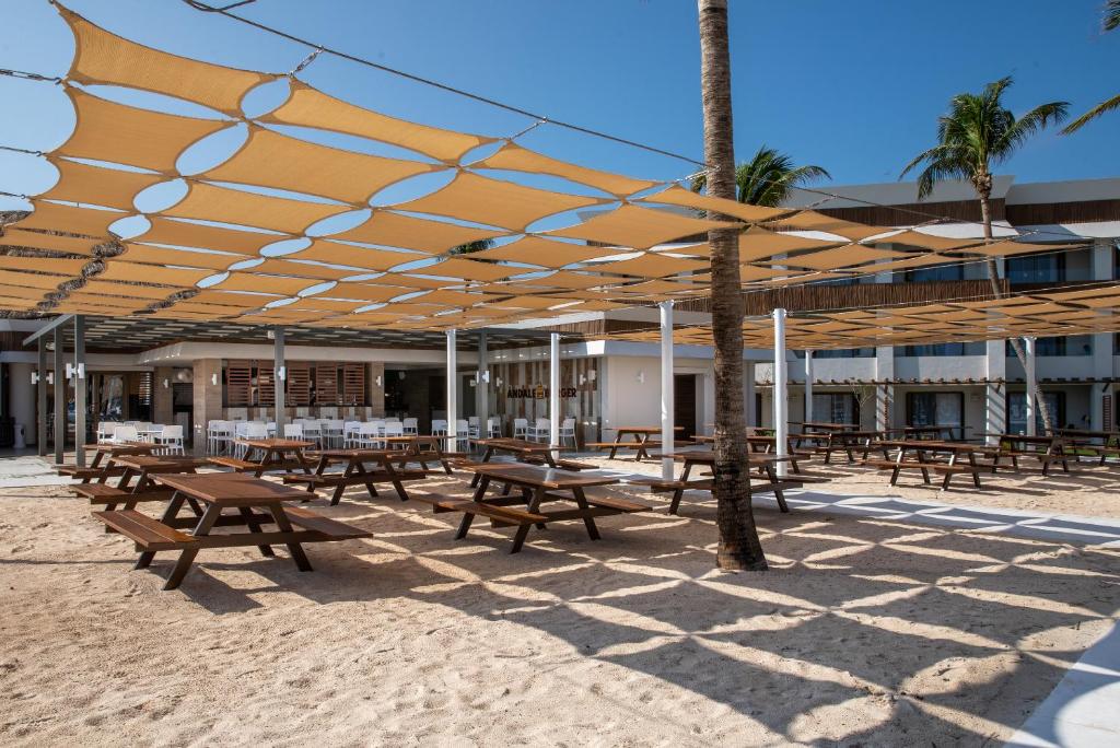 Atvērta restorāna terase ar koka galdiem, kas apklāta ar saules aizsargājošiem audumiem, apkārt palmām un gleznainai ainavai. Ideāla vieta relaksācijai pie jūras.