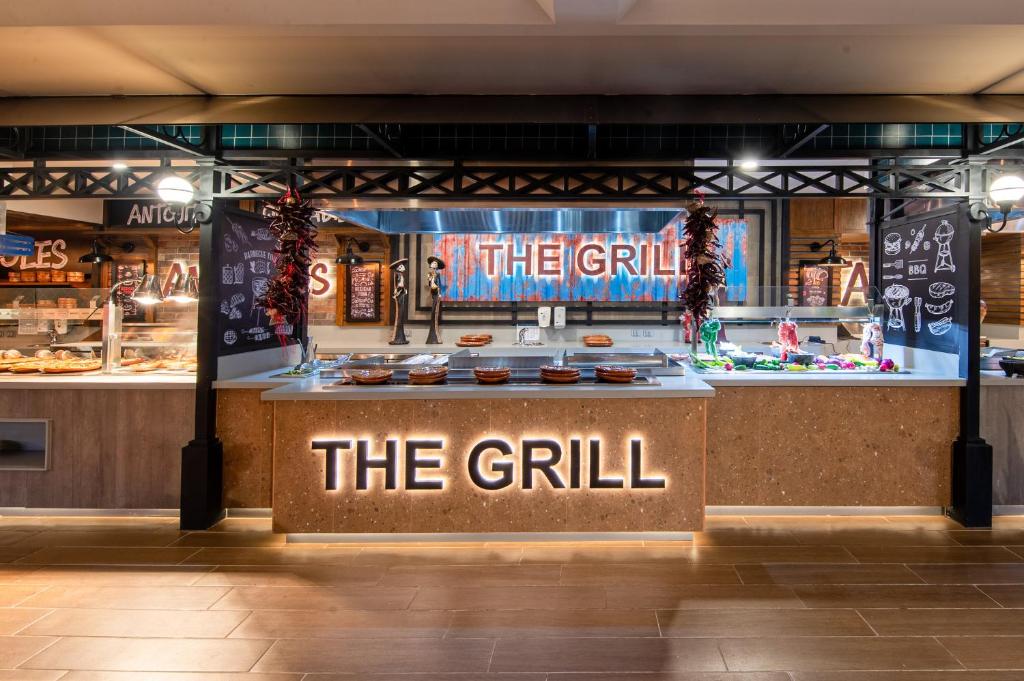 Grila restorāna zona ar modernu dizainu, izceļot nosaukumu "THE GRILL" apgaismojumā. Attēlā redzama ēdienu sagatavošanas stacija un dažādi ēdieni, kas izkārtoti uz galda.