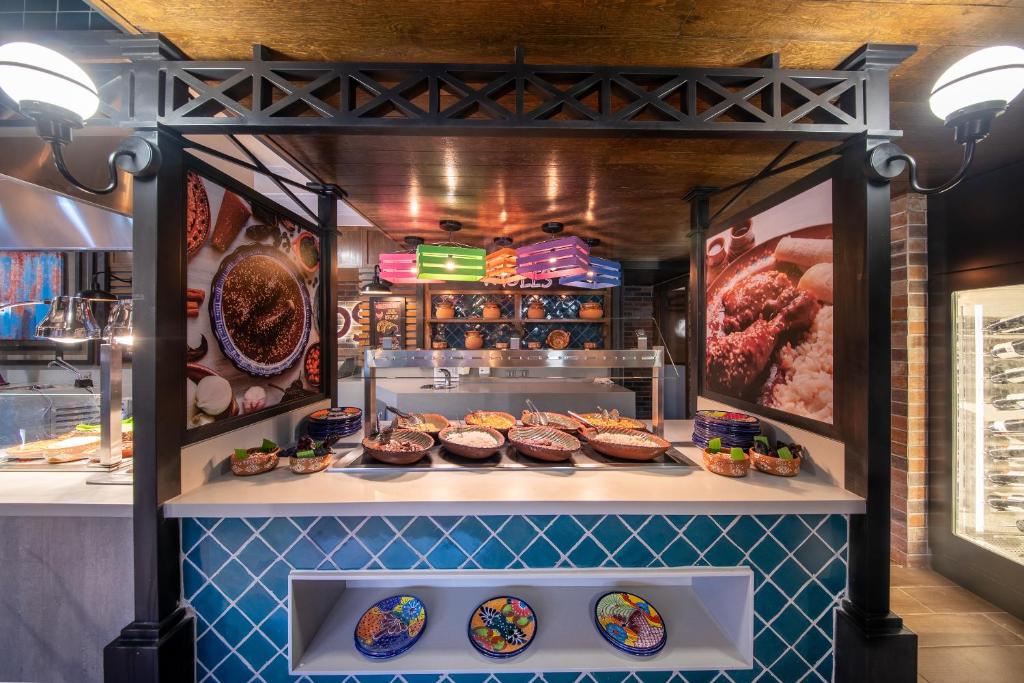 Restorāna bufete ar dažādiem ēdieniem, dekorēta ar košām plāksnēm un apgaismojumu. Fons ietver attēlus ar tradicionāliem ēdieniem un interjera elementiem.