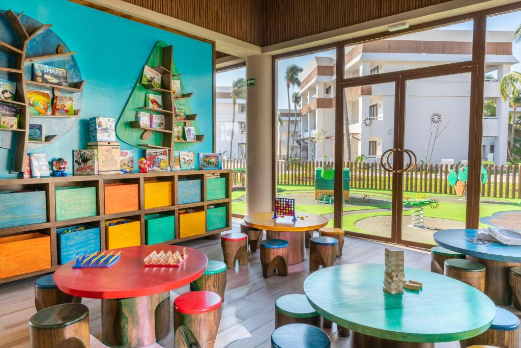 Krāsains bērnu rotaļu istabas interjers ar grāmatām plauktos, dažādām spēlēm uz galdiem un skatlogu, caur kuru redzams dārzs.