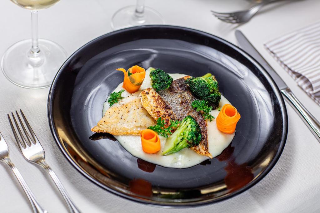 Gards ēdiens ar ceptu zivi, brokoļiem un burkānu rotājumiem, pasniegts melnā šķīvī uz baltas galdautas.