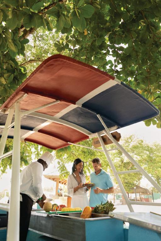 Pāris izbauda maltīti zem zaļām koku lapām, kamēr šefpavārs gatavo ēdienus pie pludmales bāra. Gaisotne ir relaksējoša un patīkama, ideāli piemērota brīvā laika pavadīšanai.