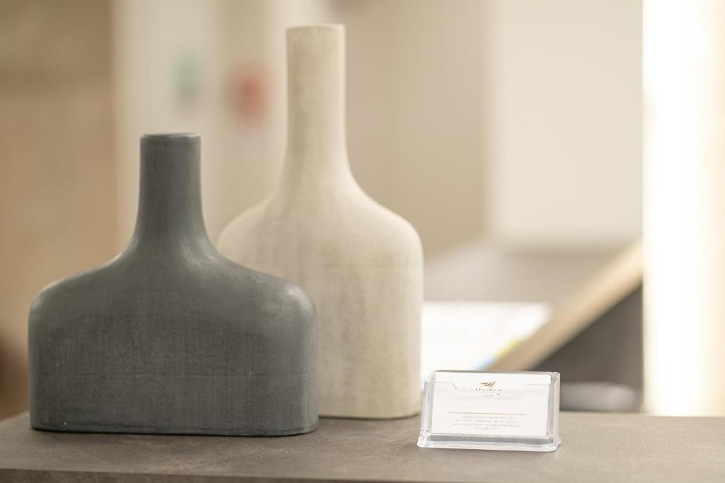 Divi moderni dekoratīvi trauki, viens pelēks un otrs balts, novietoti uz galda blakus caurspīdīgai vizītkartes turētājai.