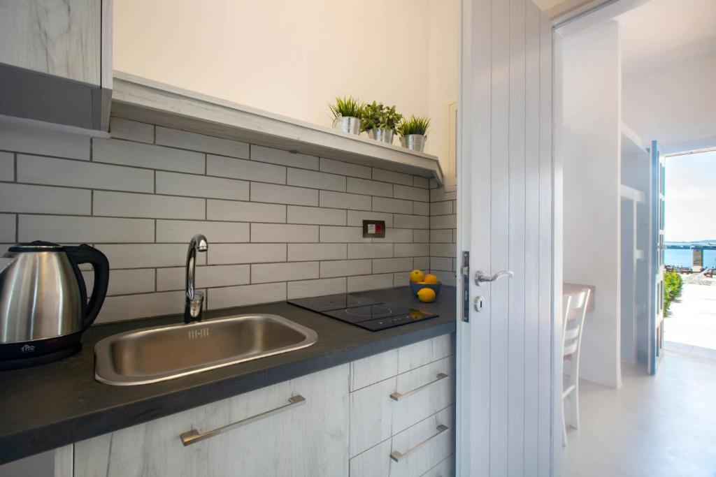 Mūsdienīga virtuves telpa ar pelēku flīžu sienu, metāla izlietni un elektrisko tējkannu. Logu durvis ved uz ārējo terasi ar skatu uz jūru.