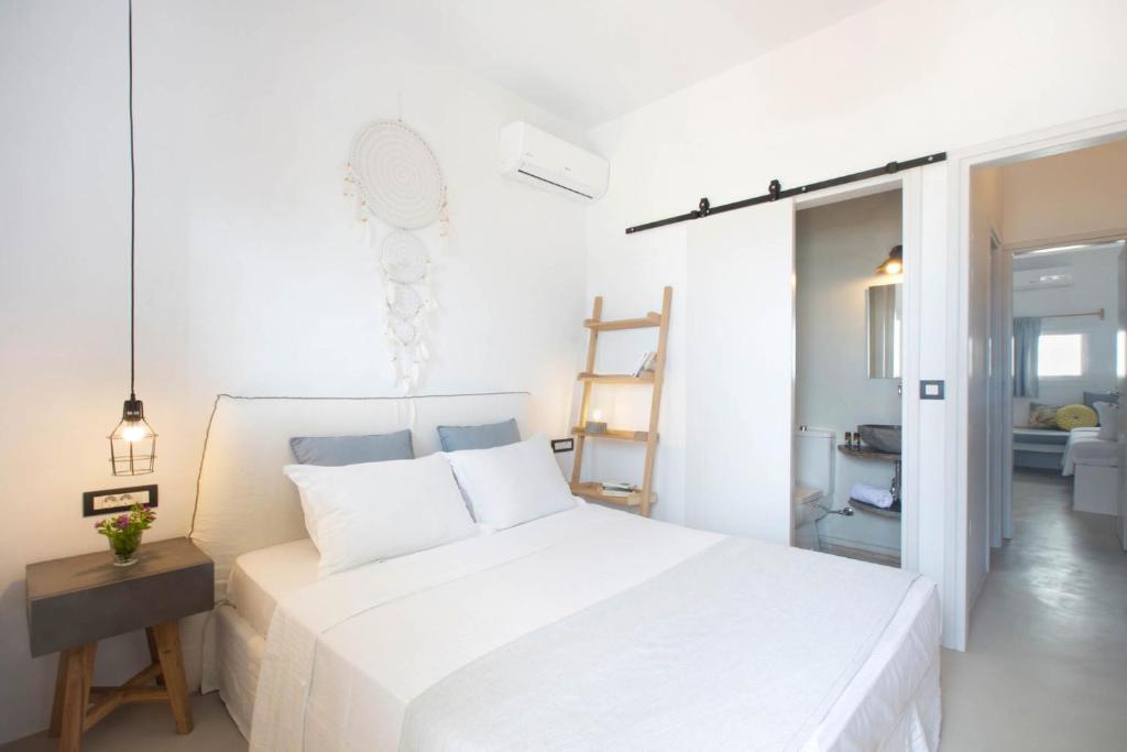 Mūsdienīga guļamistaba ar baltām sienām, ērtu gultu un dekoratīviem elementiem, piemēram, piekaramo lampu un ziedu pušķi uz naktsgaldiņa. Redzama arī piekļuve vannas istabai.