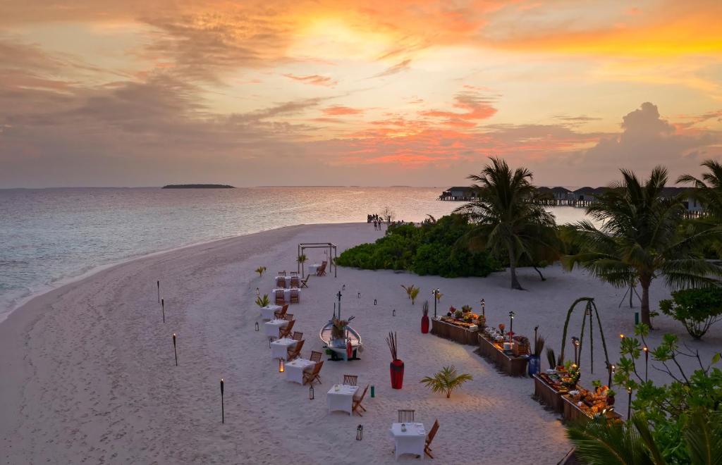 Romantiska pludmales vakariņas saulrieta gaismā, ar galdiem un krēslām smiltīs, apkārt palmas un mierīga jūra. Ideāla vieta romantiskām svinībām vai relaksējošai atpūtai.