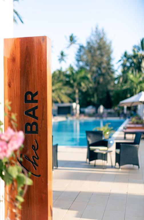 Koka stends ar uzrakstu "The BAR" blakus peldbaseinam, ko ieskauj zaļi augi un saule. Peldbaseins ir skaidrs, un ap to ir ērtas sēdvietas.