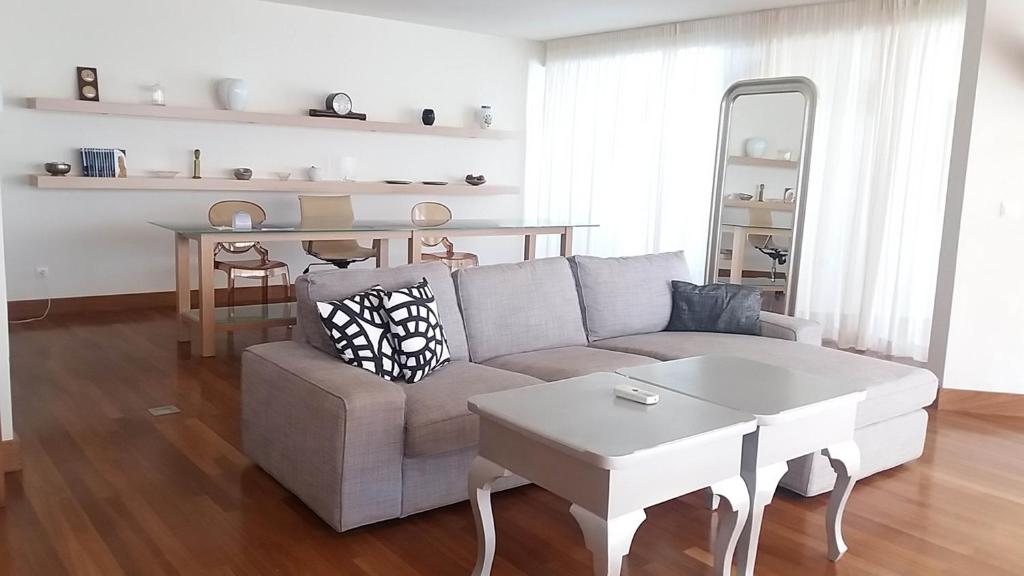 Moderns viesistabas interjers ar pelēku dīvānu, koka grīdu un elegantu galdu. Telpa ir gaiša un gaumīgi iekārtota, radot mājīgu atmosfēru.