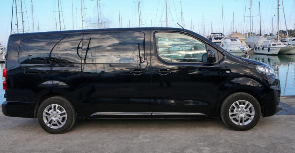 Melns furgons ar modernu dizainu, izvietots ostā starp kuģiem. Ideāls transportlīdzeklis ģimenes vai grupu ceļojumiem.