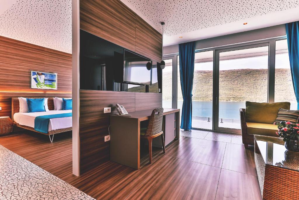 Mūsdienīga viesnīcas numura interjers ar koka apdari, divguļamo gultu un viesistabu ar skatu uz ūdeni. Numurā ir arī televizors, rakstāmgalds un dīvāns ar puķu kompozīciju.