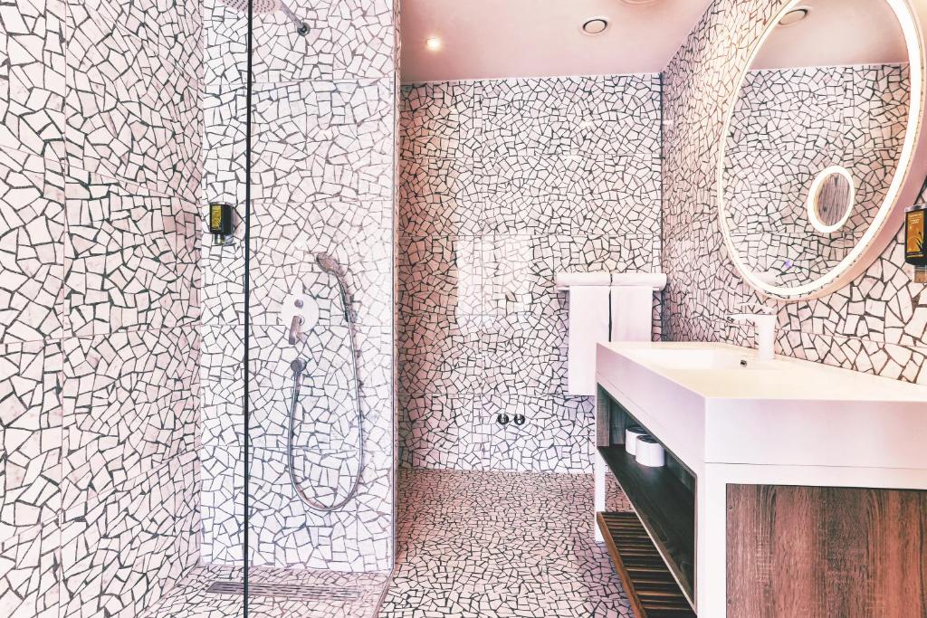 Mūsdienīgs vannas istabas interjers ar mozaīkas dizainu, kas ietver dušu, izlietni un apaļu spoguli. Vannas istaba izceļas ar gaišām krāsām un moderniem elementiem.