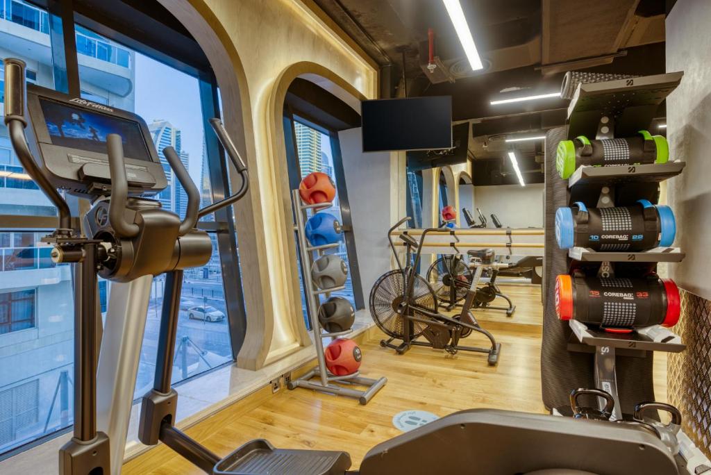 Mūsdienīgs sporta zāles interjers ar dažādiem trenažieriem un svaru stendiem, izceļot koka grīdu un lieliem logiem, kas nodrošina dabīgu apgaismojumu.