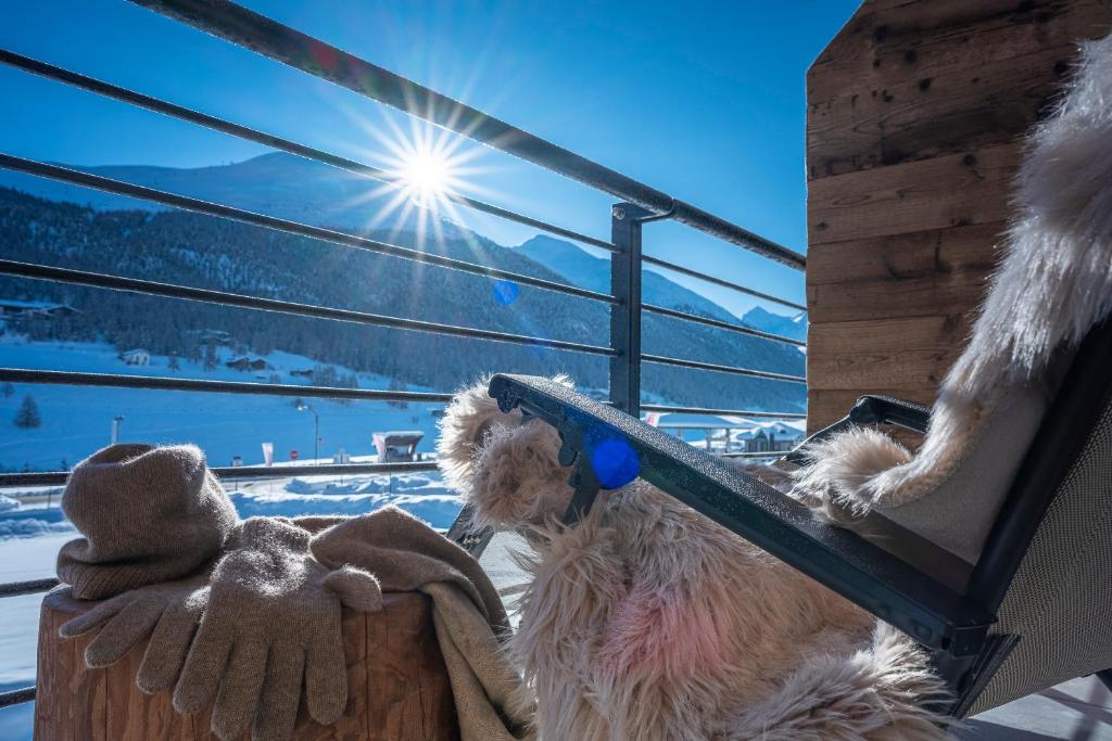 Saulaina ainava ar kalniem un sniegu, kurā redzami siltie cimdi un mīksts segums, kas novietoti uz terases krēsla. Daba piešķir mierīgu un relaksējošu atmosfēru.