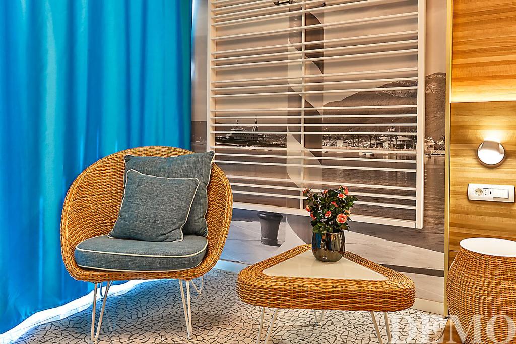 Mūsdienīgs interjers ar vītņu krēslu un mazu galdiņu, fonā redzami zili aizkari un ainava ar jūras skatu. Piemērots relaksācijai un mājīgai atmosfērai.