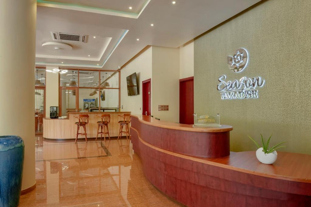 Mūsdienīgs viesnīcas reģistratūras interjers ar elegantu koka letīti un stilīgu apgaismojumu, kurā redzams "Season Paradise" zīmols.