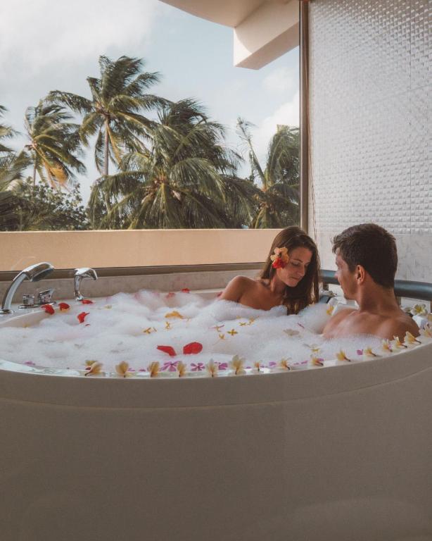 Pāris relaksējas burbuļu vannā, apkārt plūst ziedlapiņas, un loga fonā redzami palmām.