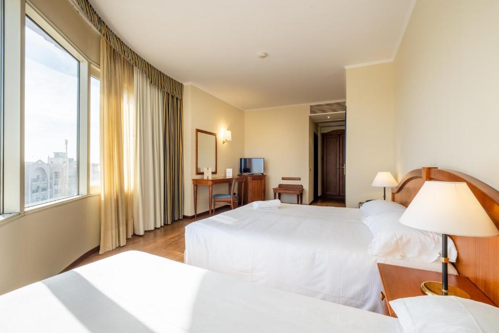 Gaiša viesnīcas numuriņa iekārtojums ar divām gultām, rakstāmgaldu, televizoru un plašiem logiem, caur kuriem ieplūst dabiska gaisma.