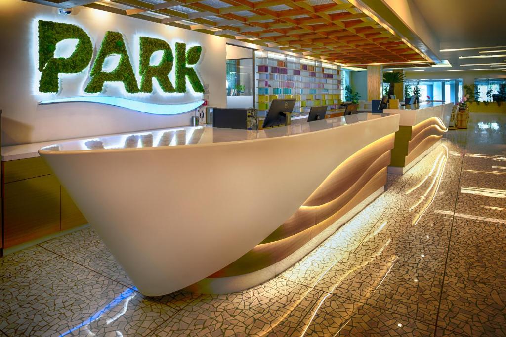 Mūsdienīgs viesnīcas reģistratūras galds ar zaļo "PARK" logo, kas izgatavots no sūnām, un apgaismojumu, kas rada patīkamu atmosfēru. Interjers ir stilīgs un krāsains, ar modernām iekārtām un augu elementiem.