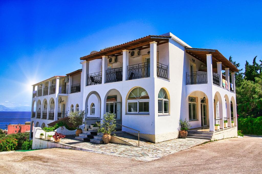 Balta villa ar arku logiem un balkoniem, kas atrodas kalnā ar skatu uz jūru. Ap to ir zaļumi un puķu podi, radot mierīgu un pievilcīgu atmosfēru.