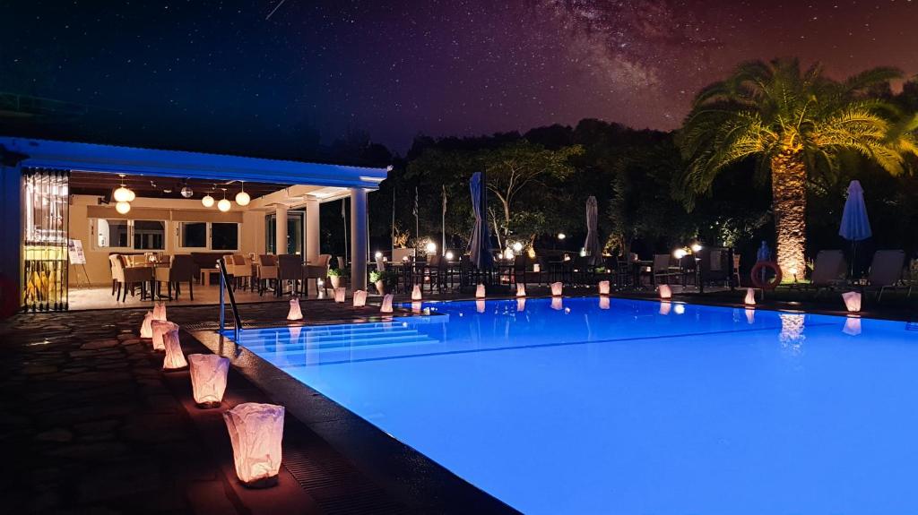 Nakts ainava ar zilo peldbaseinu, apgaismotām lampām un palmām, kas rada relaksējošu atmosfēru. Blakus baseinam ir restorāns ar galdiem un krēsliem.