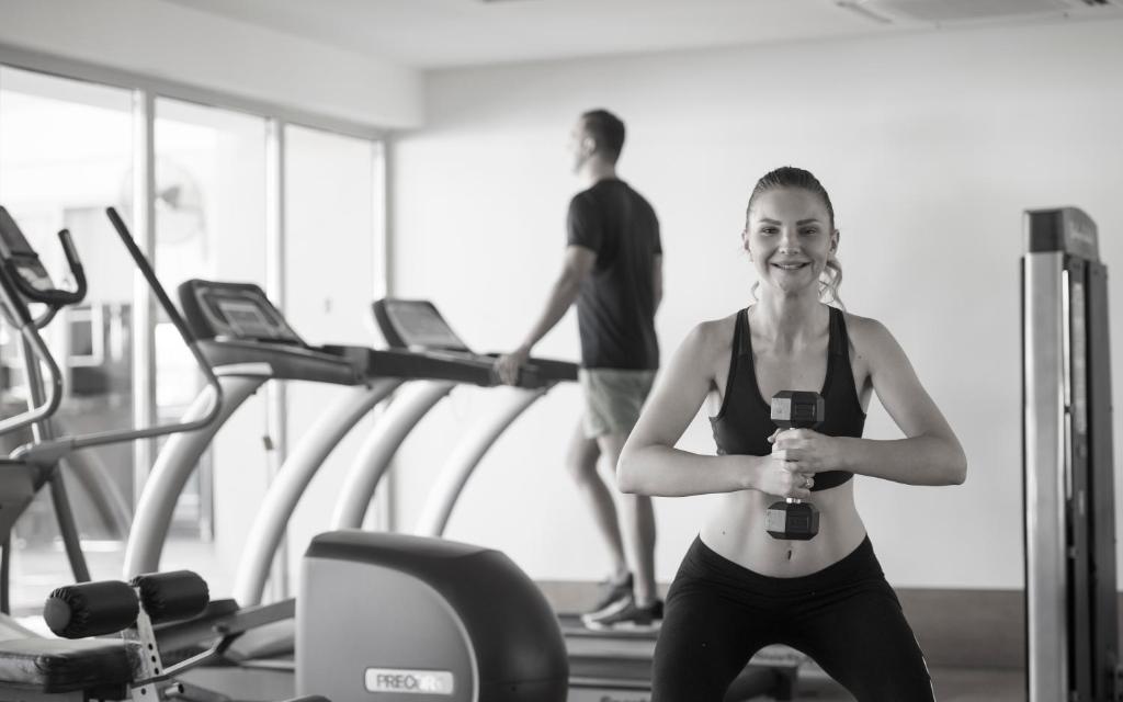 Sieviete sporta zālē tur rokās hanteles, kamēr vīrietis uz skrejceliņa veic treniņu. Ideāla aina veselīga dzīvesveida veicināšanai.