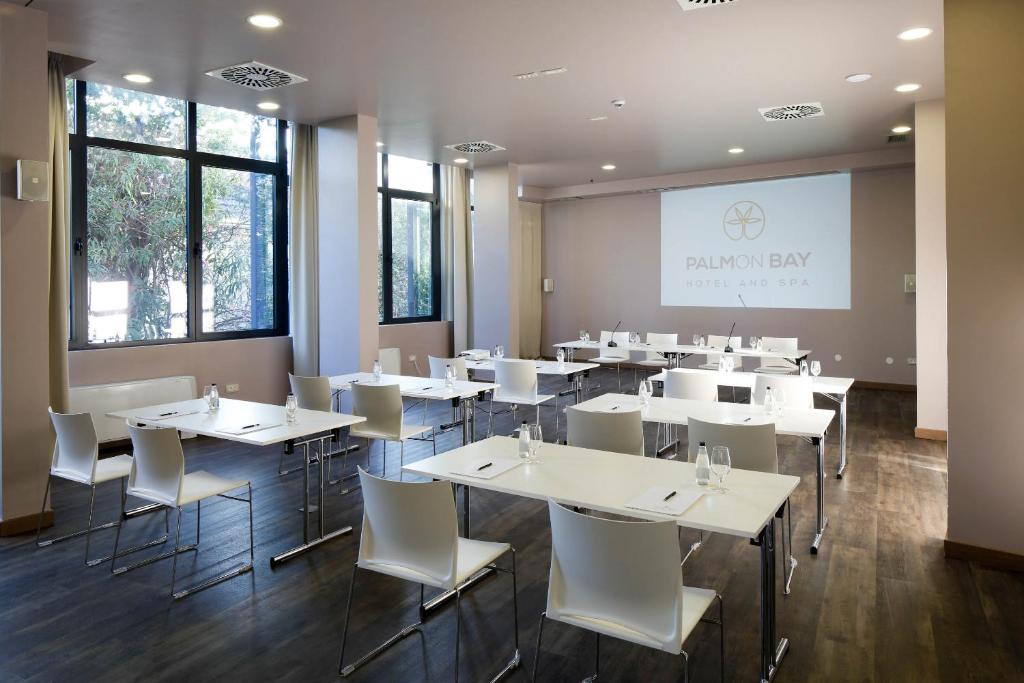 Mūsdienīgs konferenču telpas interjers ar lieliem logiem un dabīgu apgaismojumu, kurā ir sakārtotas baltas galdi un krēsli. Telpā redzama Palmon Bay viesnīcas un SPA logo.
