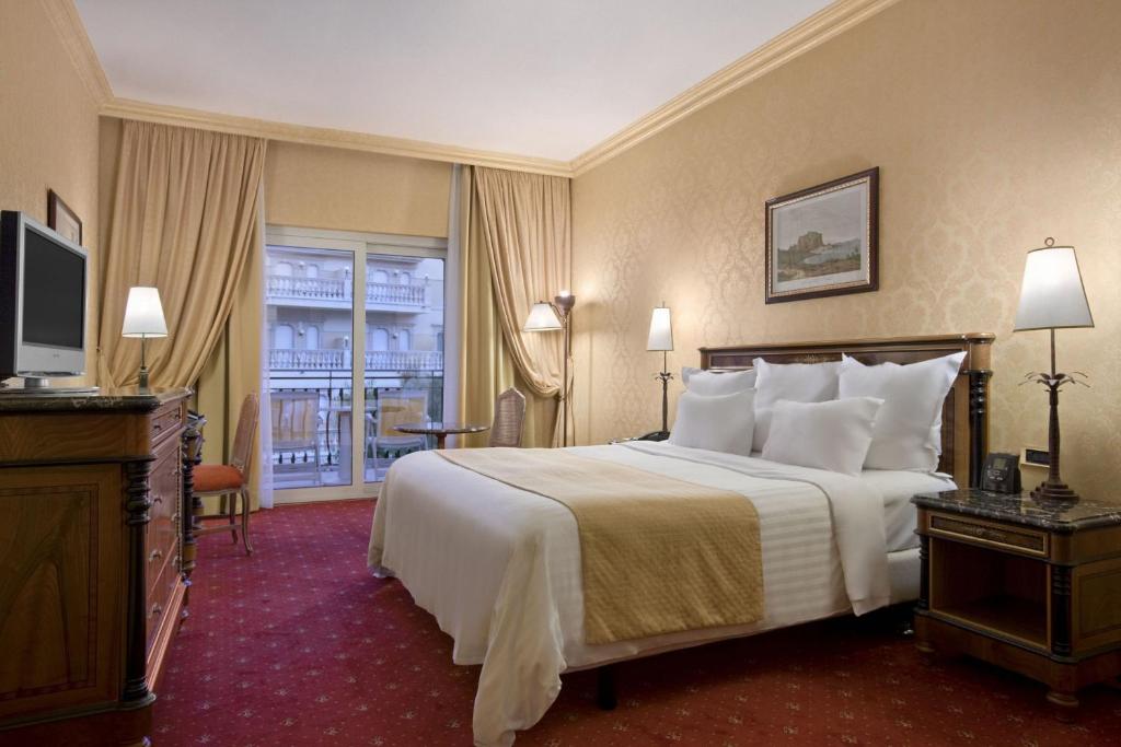 Eleganta viesnīcas numura iekārtojums ar plašu gultu, mīkstām spilveniem un stilīgu mēbelējumu. Logā redzama balkona skats, radot patīkamu atmosfēru.