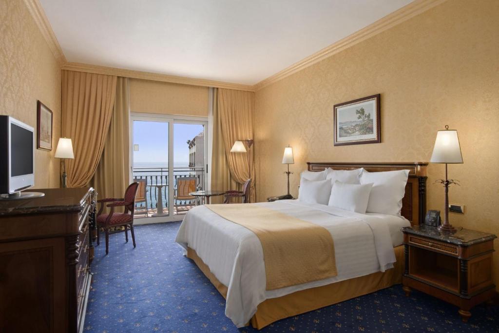 Eleganta viesnīcas numura interjers ar divguļamo gultu, dīvānu un balkonu, no kura paveras skats uz jūru. Telpā ir televizors un komfortabli mēbeles.