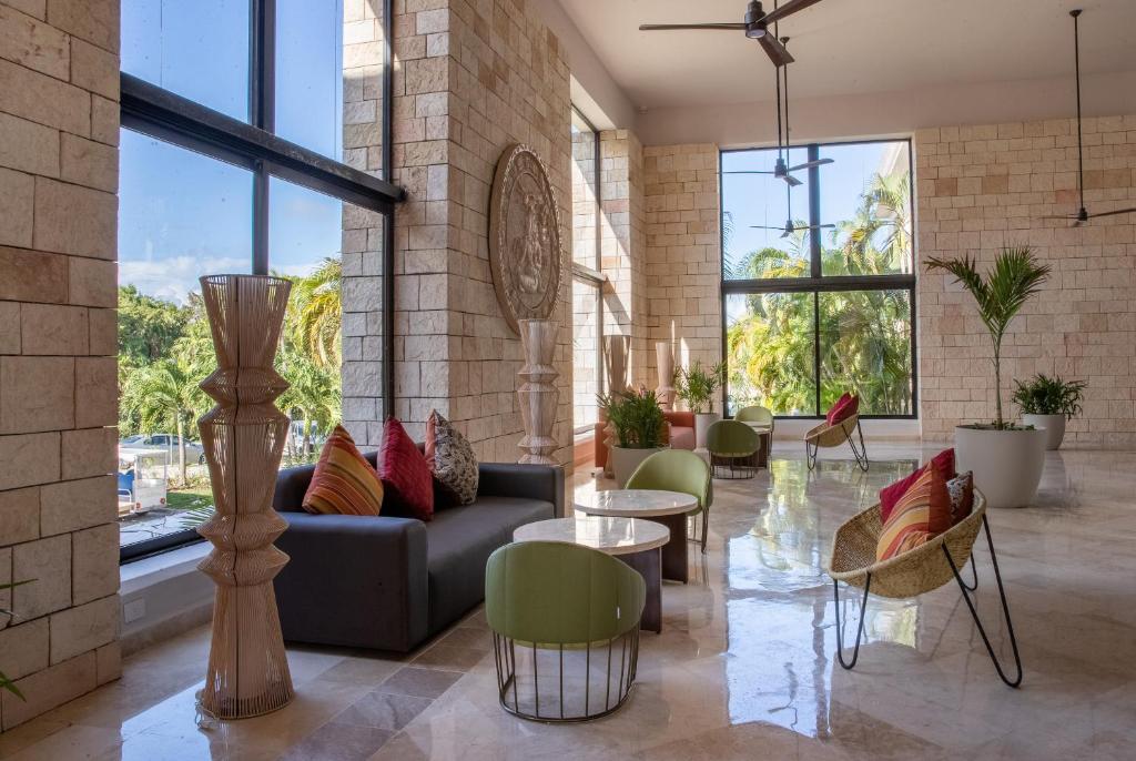 Mūsdienīgs interjers ar ērtiem dīvāniem un galdiem, kas izvietoti plašā telpā ar lieliem logiem un dabīgu apgaismojumu. Telpā redzami zaļie augi un dekoratīvi elementi, radot relaksējošu atmosfēru.