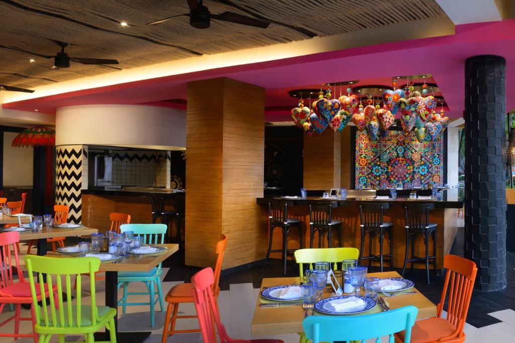 Krāsains restorāna interjers ar spilgtiem krēsliem un dekoratīvām lampām, radot dinamisku atmosfēru. Sienas rotā krāsainas mozaīkas un stilīgas akcentu detaļas.