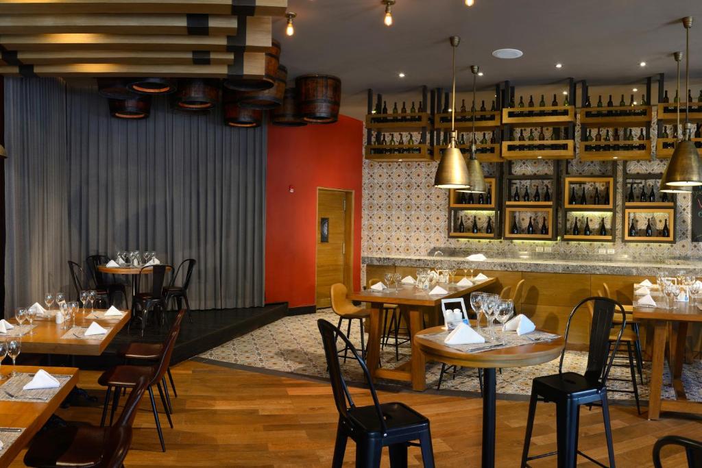 Mūsdienīgs restorāna interjers ar koka mēbelēm un īpaši izstrādātu apgaismojumu. Uz sienām ir redzami rāmji ar pudelēm un dekoratīvi elementi, kas veido siltu atmosfēru.