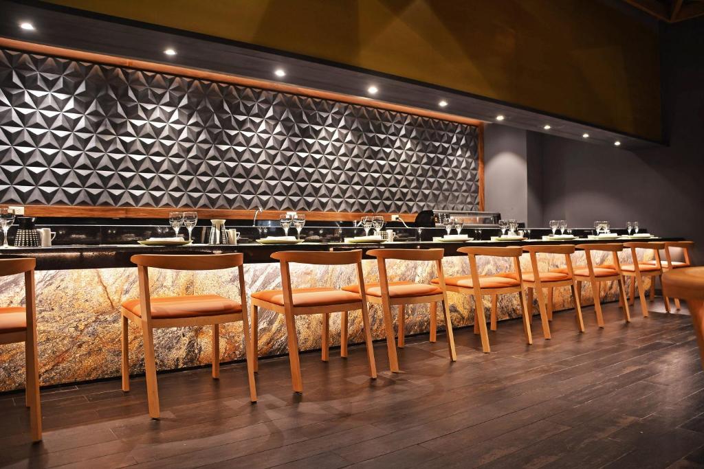Mūsdienīga restorāna interjers ar elegantu apgaismojumu un stilīgām koka krēsliem, kas izvietoti pie garas galda virsmas. Fona siena ar ģeometrisku reljefu piešķir telpai unikālu raksturu.
