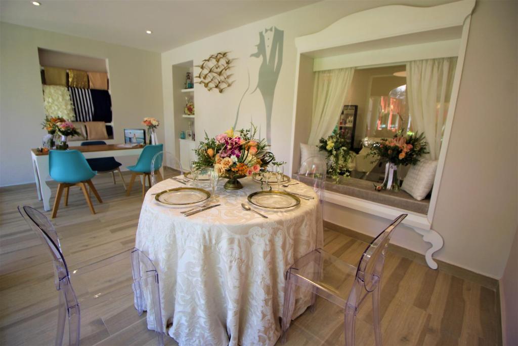 Eleganta ēdamzona ar apaļu galdu, dekorētu ar ziediem un sudraba traukiem. Interjers ir moderns ar gaišām krāsām un caurspīdīgām krēslām.