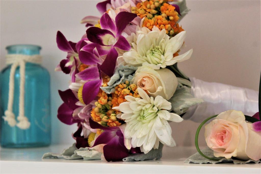 Krāsains kāzu pušķis ar orhidejām, kumelītēm un rozēm, novietots blakus ziliem dekoratīviem traukiem.