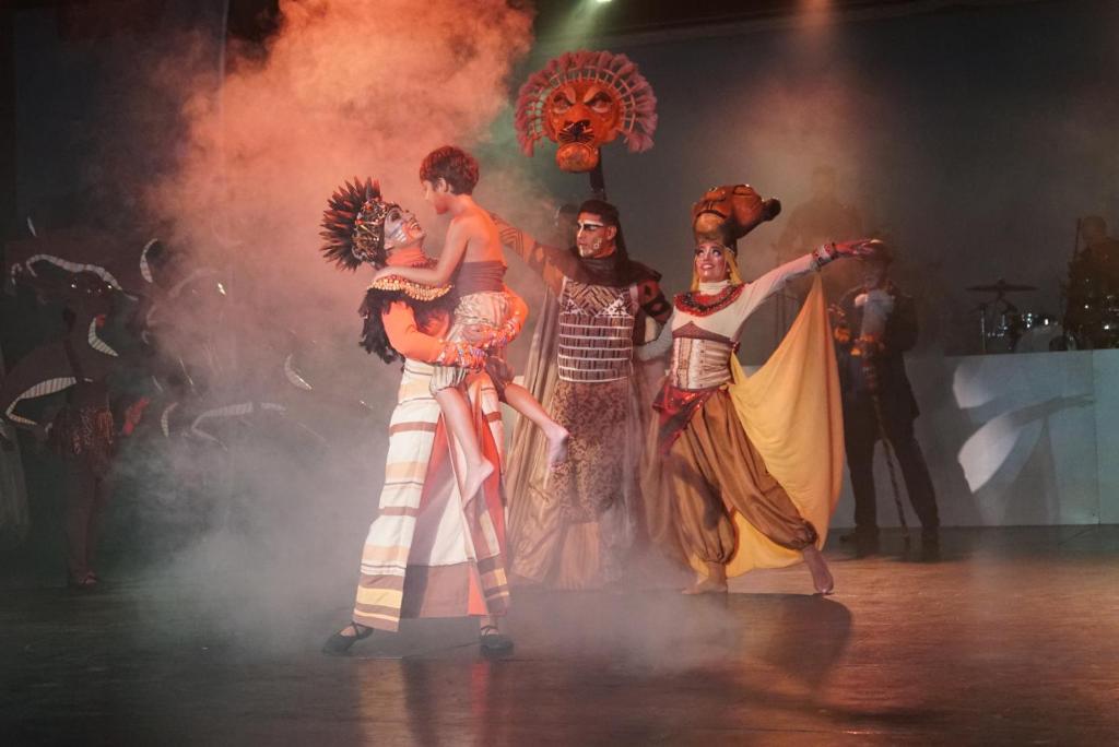 Skaistā teātra izrāde ar krāšņiem kostīmiem un dejotājiem, kas izpilda dinamiskas kustības dūmu apgaismojumā.