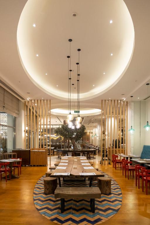 Mūsdienīgs restorāna interjers ar koka mēbelēm, izsmalcinātām gaismām un atvērtu plānojumu, kur centrā ir liels galds un dekoratīvs koki. Siltas krāsas un mūsdienīgs dizains rada patīkamu atmosfēru.