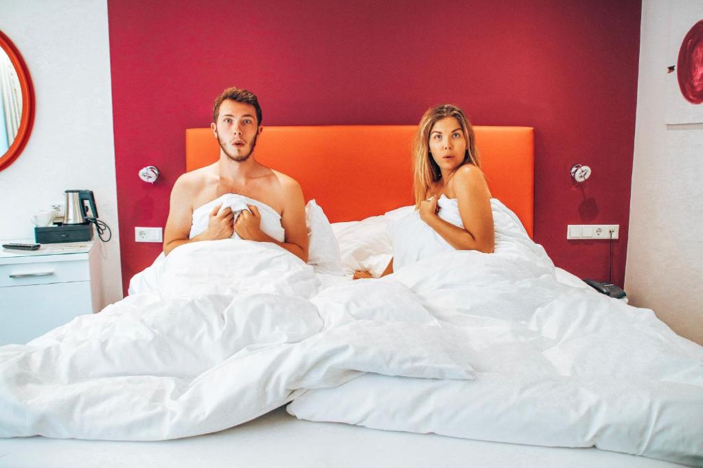 Pāris gultā ar pārsteigtām sejām, pārklāti ar baltām gultas segām. Fons ir ar sarkanu sienu un gultas galvgali.