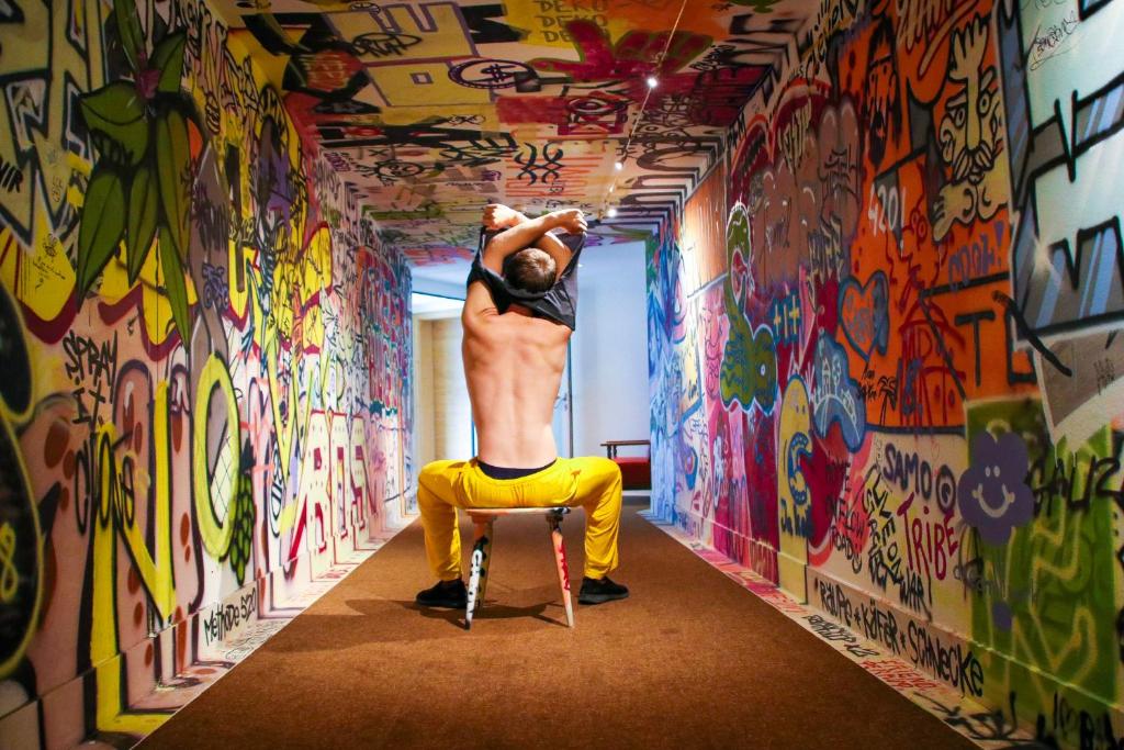 Dejotājs veic izpildījumu, sēžot uz krēsla krāsainā graffiti apdarinātā koridorā. Fons ir piepildīts ar dzīvīgu mākslu un uzrakstiem, kas pievērš uzmanību.