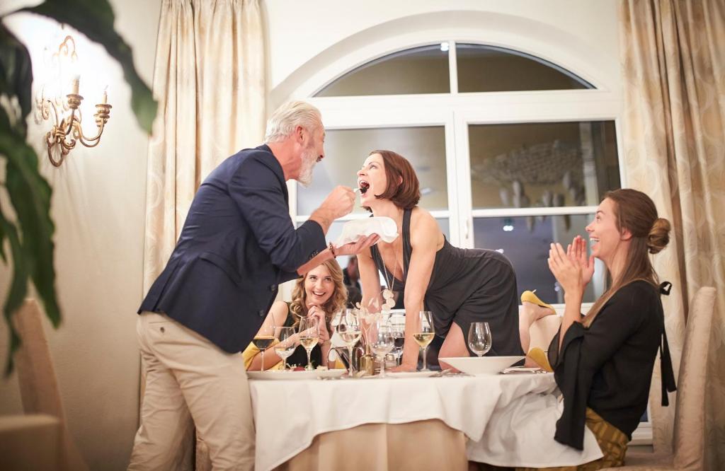 Grezns vakariņu brīdis, kurā cilvēki izbauda jautru un dzīvīgu atmosfēru, ar vienu sievieti, kas izceļas ar izteiksmīgu pozu uz galda. Slinko gaisma un eleganta apdare papildina svinību noskaņu.