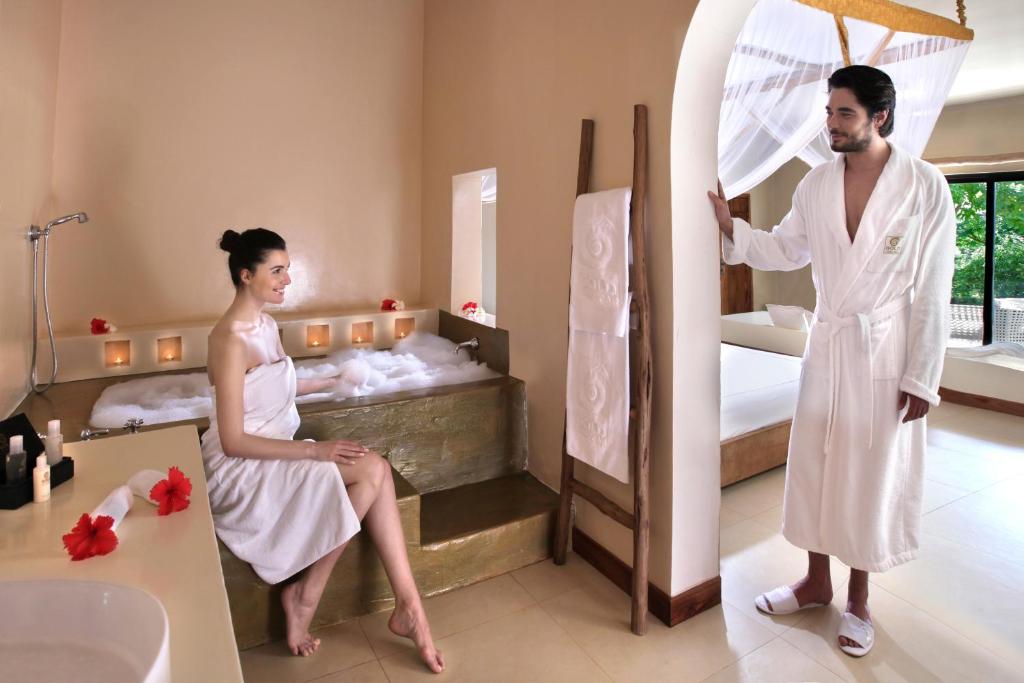 Mūsdienīgs spa interjers ar diviem cilvēkiem. Sieviete relaksējas vannā ar putām, bet vīrietis stāv durvīs, ģērbies baltā halātā. Spa atmosfēra ir mierīga un romantiska, ar svecēm un ziediem.