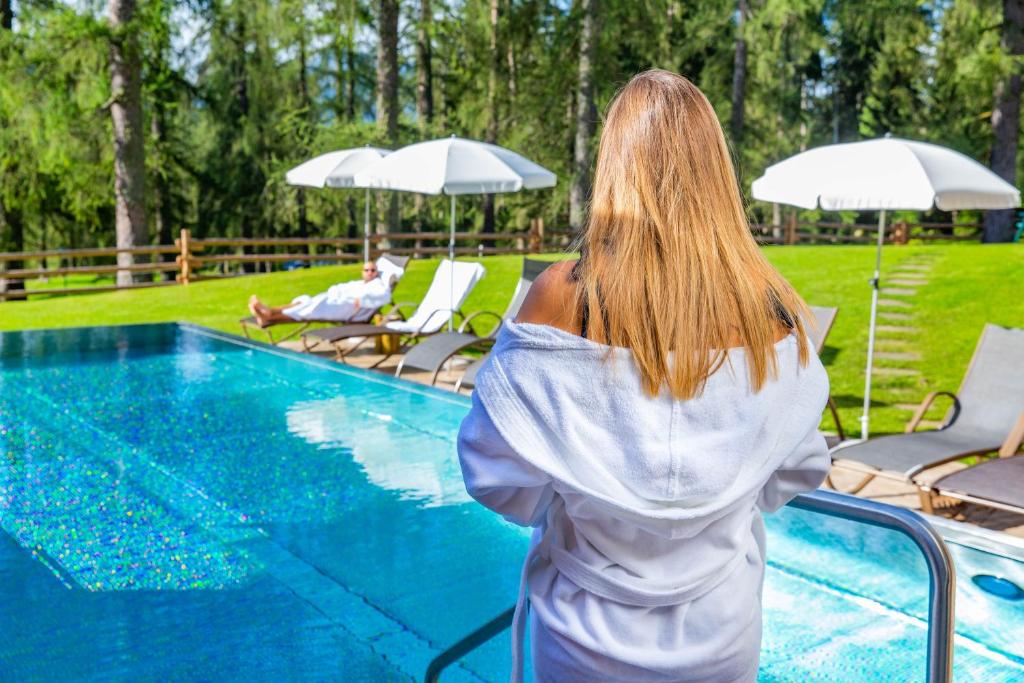 Relaksējoša ainava pie baseina ar saulē mirdzošu ūdeni un cilvēku, kas atpūšas, valkājot baltu peldmētelīti. Fons ar zaļām zālēm un koku rindām rada mierīgu atmosfēru.